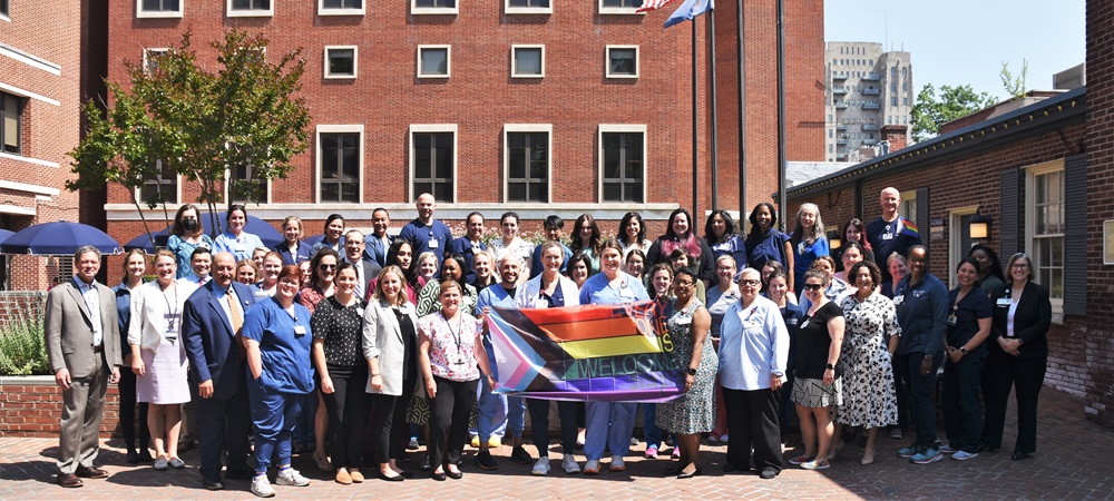 A crowd of Pennsylvania Hospital employees stand outside in the Elm Garden holding a Pride flag that says Everyone is welcome here